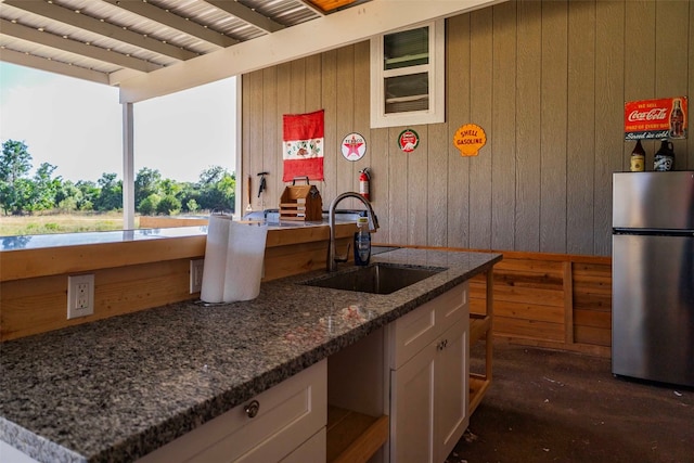 kitchen with wood walls, stainless steel refrigerator, sink, white cabinetry, and dark stone countertops