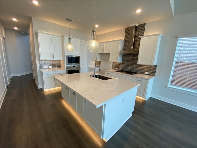 kitchen featuring wall chimney range hood, sink, an island with sink, white cabinets, and appliances with stainless steel finishes