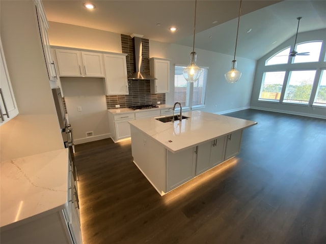 kitchen featuring wall chimney range hood, decorative light fixtures, a center island with sink, light stone counters, and white cabinets