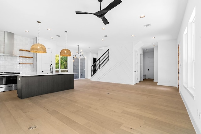 kitchen featuring light wood-type flooring, wall chimney exhaust hood, stainless steel range, a kitchen island with sink, and sink