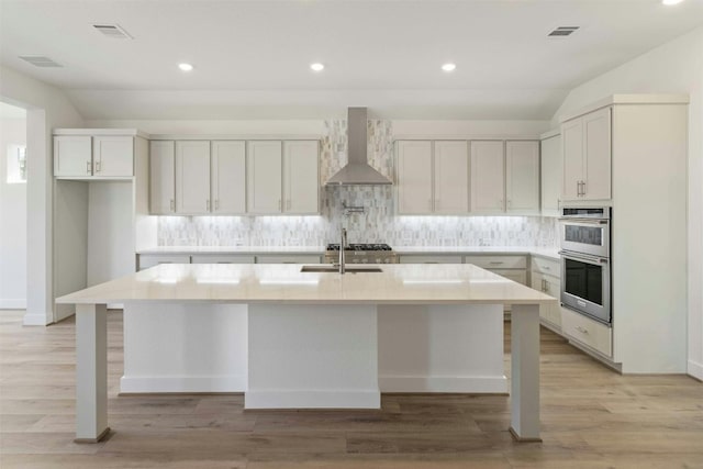 kitchen featuring wall chimney range hood, decorative backsplash, stainless steel double oven, and an island with sink