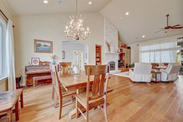 dining room with a stone fireplace, high vaulted ceiling, ceiling fan with notable chandelier, and light wood-type flooring