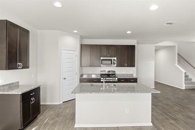 kitchen with wood-type flooring, a center island with sink, appliances with stainless steel finishes, and light stone counters