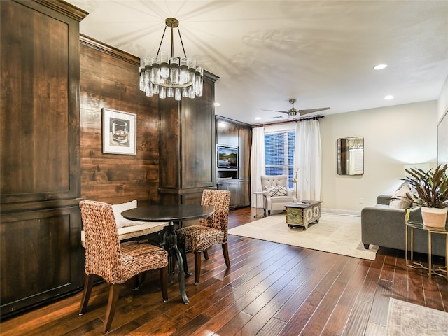 dining space with dark wood-type flooring, ceiling fan with notable chandelier, and wooden walls