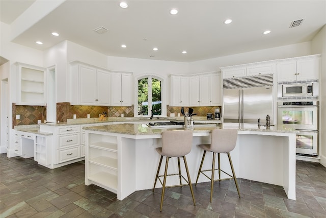 kitchen with white cabinets, backsplash, a center island with sink, and built in appliances