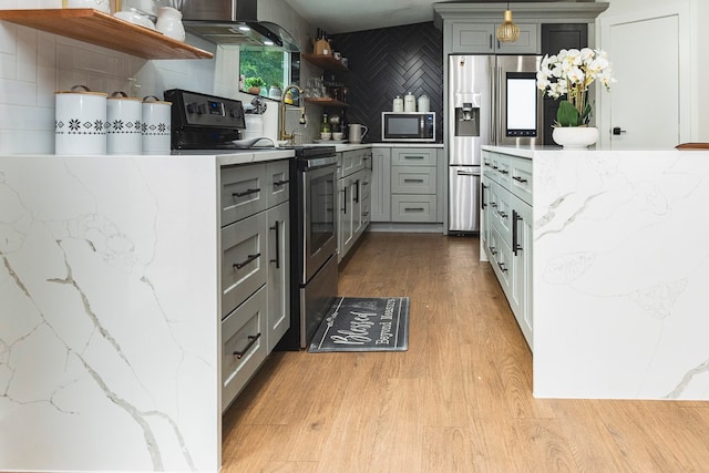 kitchen featuring gray cabinetry, backsplash, stainless steel appliances, wall chimney range hood, and light wood-type flooring