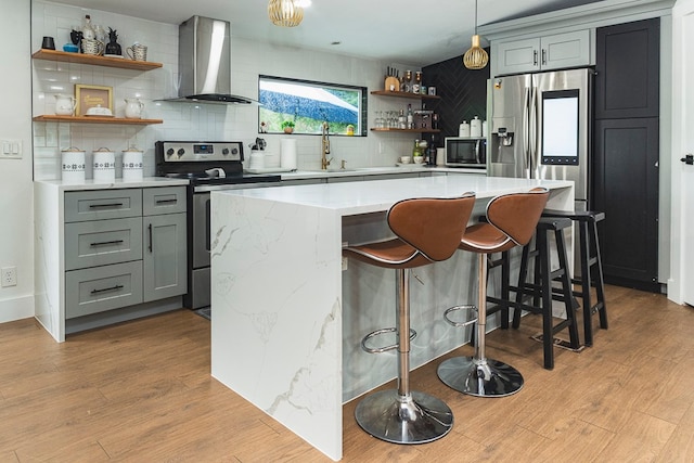 kitchen featuring appliances with stainless steel finishes, light wood-type flooring, wall chimney range hood, tasteful backsplash, and a breakfast bar area