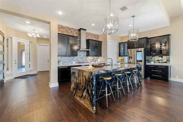 kitchen with a breakfast bar, visible vents, wall chimney range hood, an island with sink, and light stone countertops