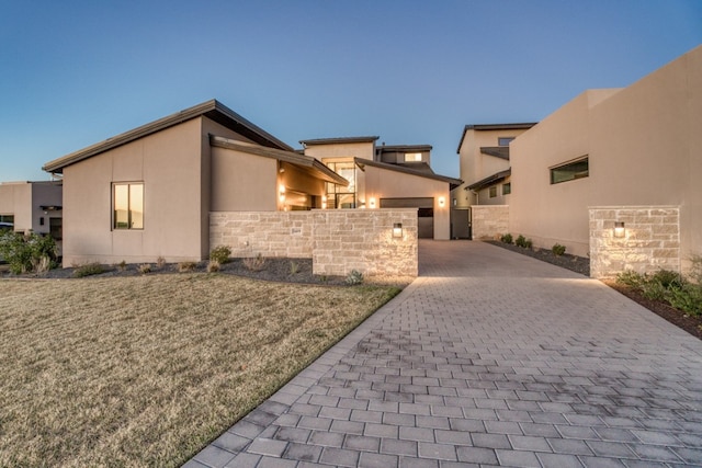 property exterior at dusk with stone siding, an attached garage, decorative driveway, a yard, and stucco siding