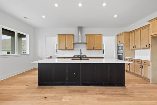 kitchen with a kitchen island with sink, wall chimney range hood, decorative backsplash, appliances with stainless steel finishes, and light hardwood / wood-style floors