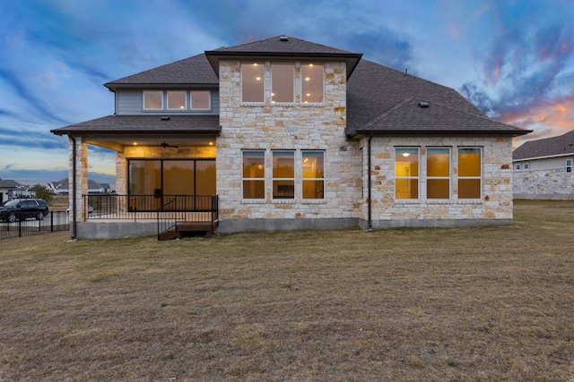 back of house at dusk featuring stone siding, a ceiling fan, a lawn, and roof with shingles