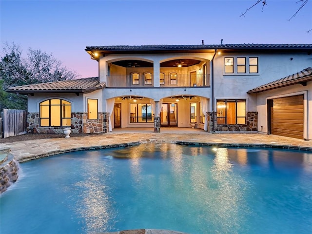 back house at dusk featuring a garage, a balcony, ceiling fan, and a patio area