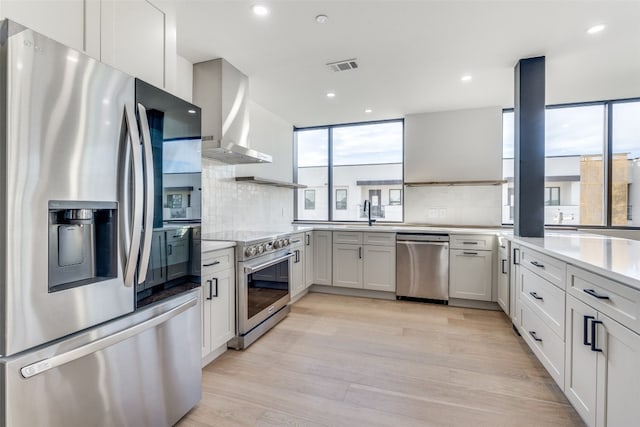 kitchen with wall chimney exhaust hood, appliances with stainless steel finishes, decorative backsplash, and light hardwood / wood-style floors