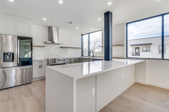 kitchen with light hardwood / wood-style flooring, backsplash, stainless steel appliances, wall chimney exhaust hood, and white cabinets