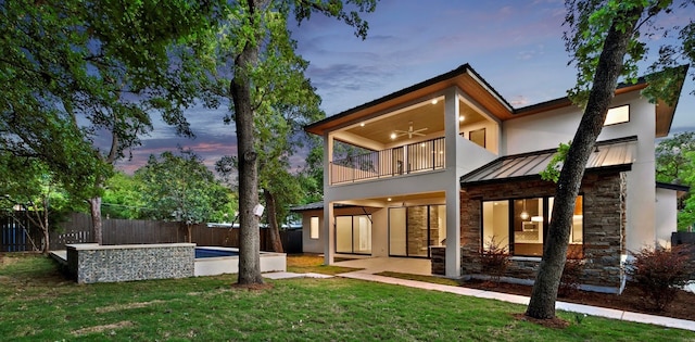 back house at dusk with a balcony, a patio area, and a yard