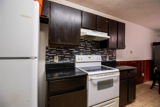 kitchen with under cabinet range hood, a wainscoted wall, light tile patterned floors, white appliances, and a textured ceiling