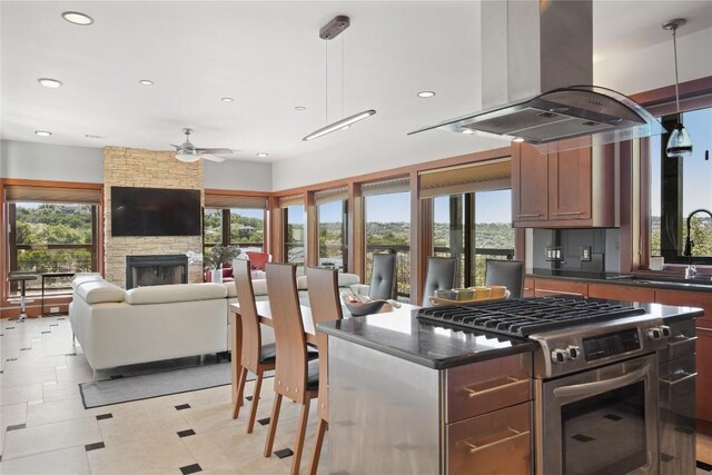 kitchen featuring ceiling fan, island exhaust hood, pendant lighting, a stone fireplace, and stainless steel gas range