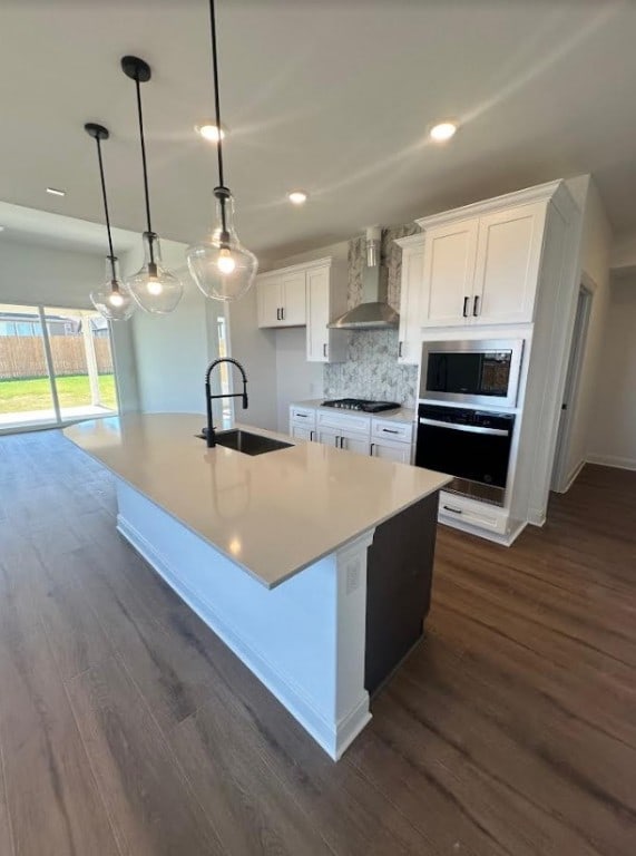 kitchen featuring pendant lighting, white cabinets, a kitchen island with sink, wall chimney range hood, and appliances with stainless steel finishes