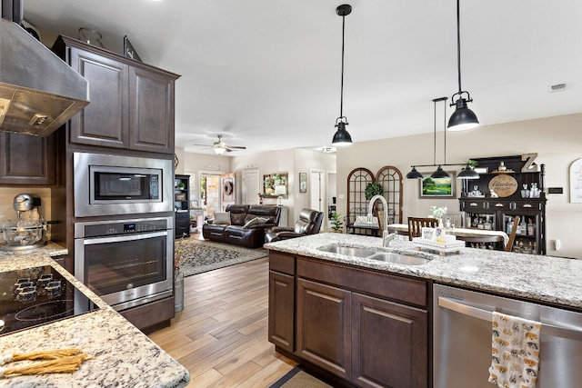 kitchen with light wood finished floors, wall chimney exhaust hood, appliances with stainless steel finishes, dark brown cabinets, and a sink