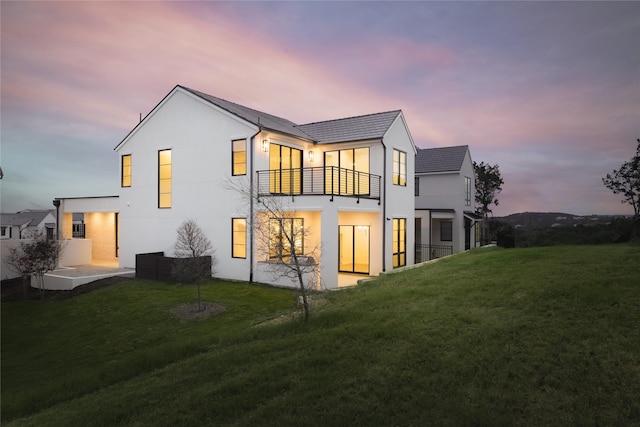 back house at dusk featuring a lawn, a patio area, and a balcony