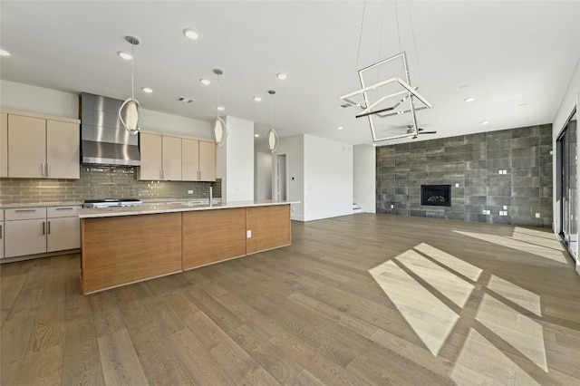 kitchen with hardwood / wood-style floors, wall chimney exhaust hood, a fireplace, an island with sink, and sink