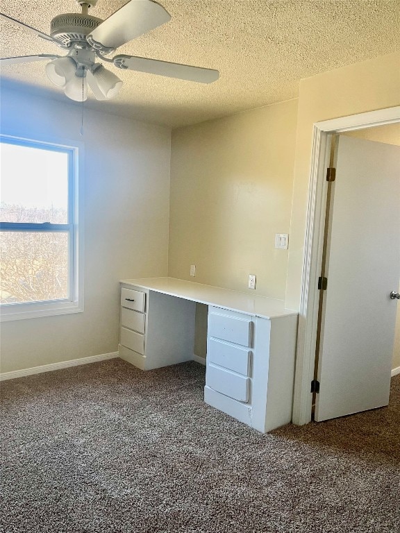 unfurnished office featuring dark colored carpet, built in desk, ceiling fan, and a textured ceiling