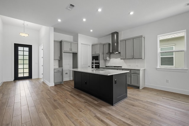 kitchen with decorative backsplash, wall chimney exhaust hood, light hardwood / wood-style floors, stainless steel microwave, and a center island with sink