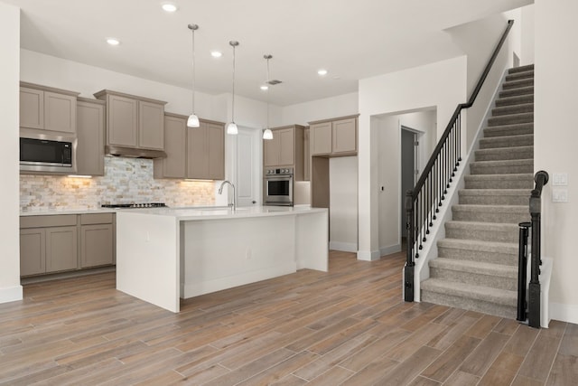 kitchen featuring backsplash, light hardwood / wood-style floors, decorative light fixtures, a center island with sink, and appliances with stainless steel finishes