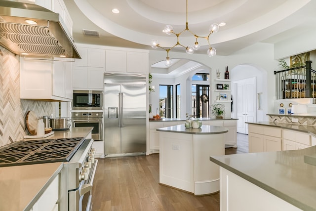 kitchen featuring island range hood, hanging light fixtures, built in appliances, a tray ceiling, and white cabinets