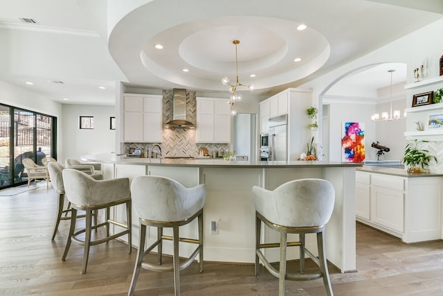 kitchen featuring white cabinets, a kitchen island with sink, a tray ceiling, and wall chimney range hood