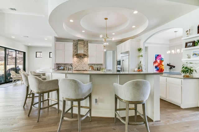 kitchen with a kitchen island with sink, wall chimney range hood, white cabinetry, and decorative light fixtures