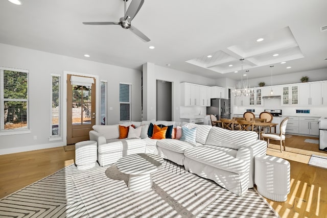 living room with ceiling fan with notable chandelier, coffered ceiling, light wood-type flooring, and a tray ceiling