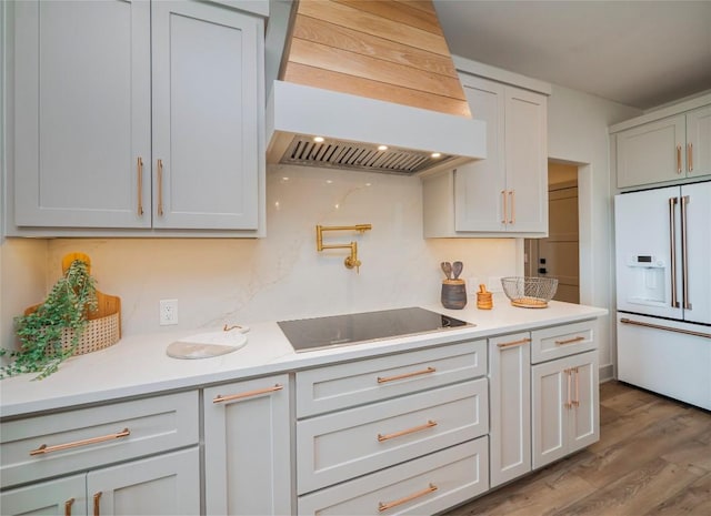 kitchen with black electric stovetop, white built in refrigerator, custom range hood, and wood-type flooring