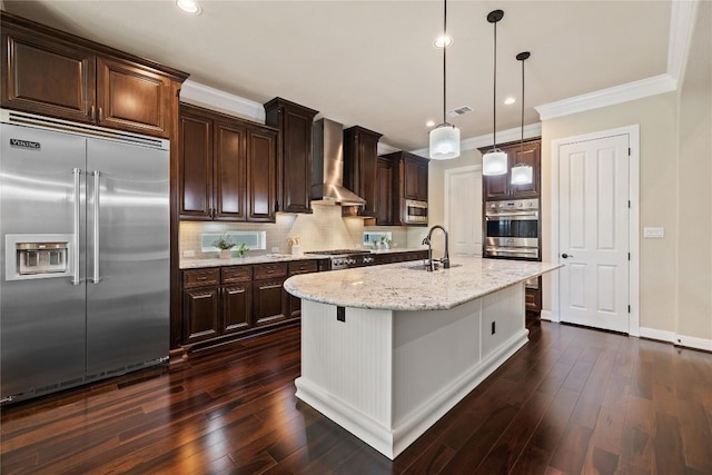 kitchen with hanging light fixtures, wall chimney range hood, backsplash, built in appliances, and a kitchen island with sink