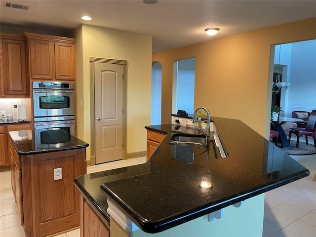 kitchen with dark stone counters, double oven, sink, an island with sink, and light tile patterned flooring