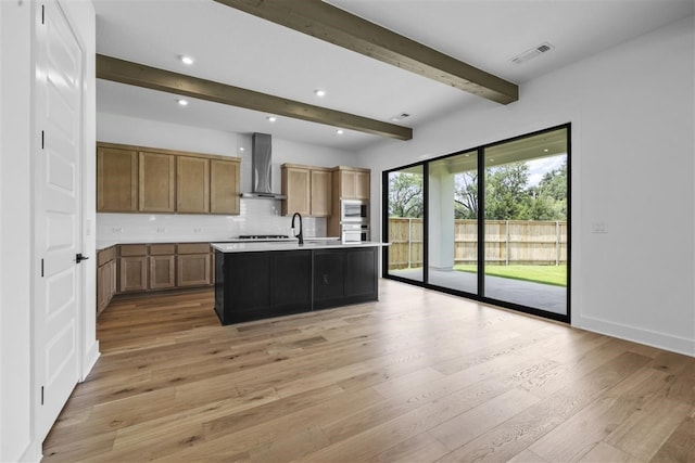 kitchen with a center island with sink, sink, wall chimney exhaust hood, beam ceiling, and light wood-type flooring