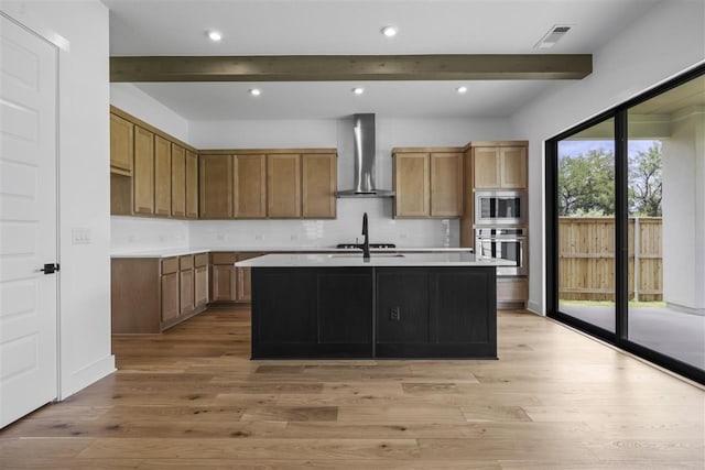 kitchen featuring stainless steel microwave, an island with sink, beamed ceiling, wall chimney range hood, and light hardwood / wood-style flooring