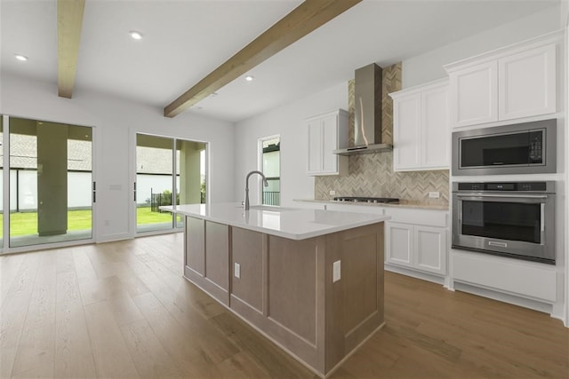 kitchen with beamed ceiling, white cabinetry, stainless steel appliances, wall chimney exhaust hood, and a center island with sink