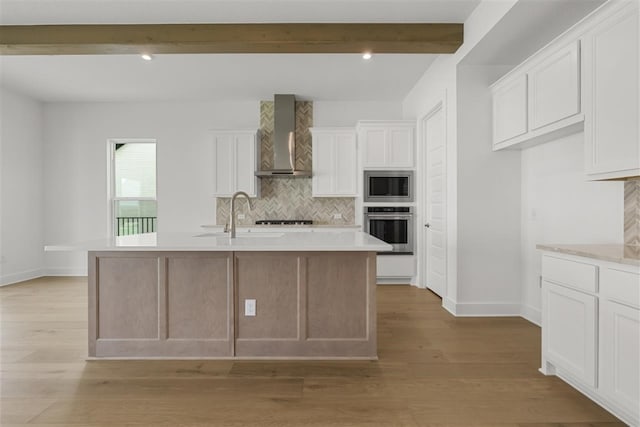 kitchen featuring appliances with stainless steel finishes, white cabinetry, wall chimney exhaust hood, and a kitchen island with sink