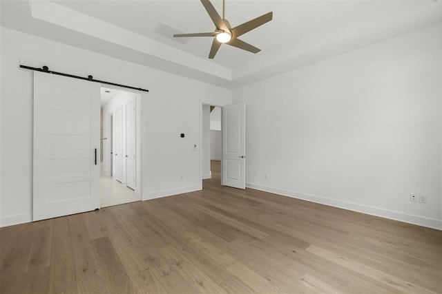 unfurnished bedroom featuring light hardwood / wood-style floors, a raised ceiling, a barn door, and ceiling fan