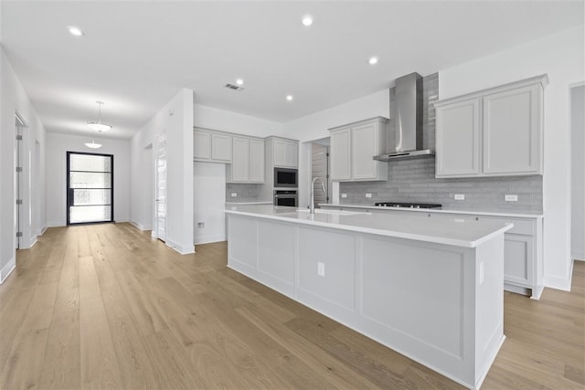 kitchen with stainless steel appliances, wall chimney range hood, sink, an island with sink, and light hardwood / wood-style flooring