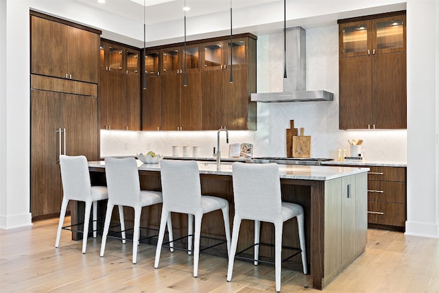 kitchen with a center island with sink, wall chimney exhaust hood, pendant lighting, and light hardwood / wood-style flooring