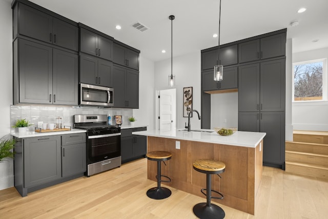 kitchen with stainless steel appliances, sink, pendant lighting, light hardwood / wood-style floors, and an island with sink