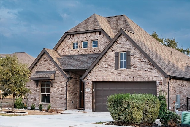 french provincial home with a garage, concrete driveway, brick siding, and roof with shingles