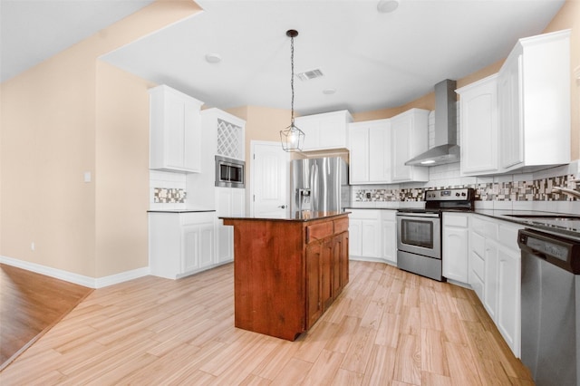 kitchen with appliances with stainless steel finishes, white cabinetry, a kitchen island, light wood-type flooring, and wall chimney exhaust hood