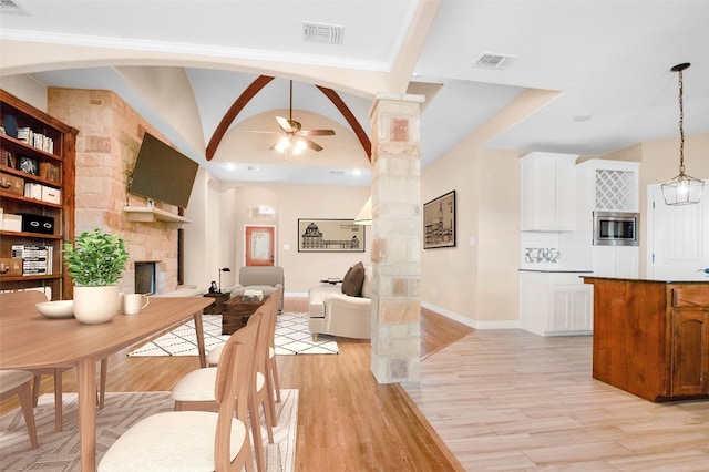 dining room with light hardwood / wood-style floors, ornate columns, ceiling fan with notable chandelier, a stone fireplace, and vaulted ceiling with beams