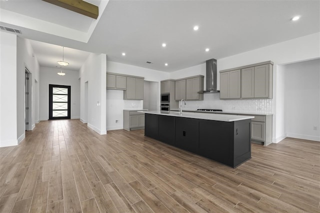 kitchen featuring light hardwood / wood-style floors, a center island with sink, decorative backsplash, wall chimney exhaust hood, and gray cabinets