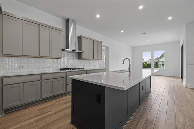 kitchen with gray cabinets, wall chimney exhaust hood, a center island with sink, and light hardwood / wood-style flooring