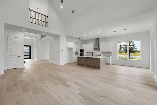 kitchen with high vaulted ceiling, a center island with sink, white cabinets, wall chimney range hood, and light wood-type flooring