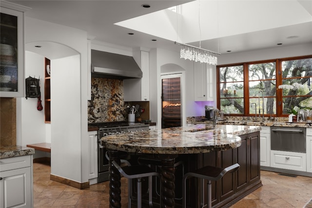 kitchen featuring white cabinets, an island with sink, high end stainless steel range oven, wall chimney exhaust hood, and a kitchen breakfast bar